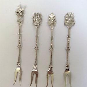 May include: Set of four silver-toned forks with decorative handles. Each handle features a different Dutch-themed design, including a windmill, a group of people, a man carrying a barrel, and a scene with a horse and cart.