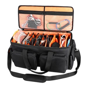 May include: Black camera bag with multiple compartments and an orange interior. The bag has a shoulder strap and two handles. The bag is filled with various cables and accessories.