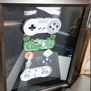 May include: A framed display of disassembled parts of a Super Nintendo controller. The controller is white with gray buttons and a green circuit board. The parts are arranged in a grid pattern on a black background.
