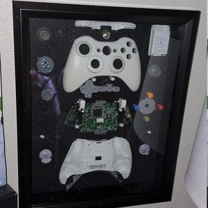 May include: A white Xbox 360 controller disassembled and displayed in a black frame. The controller is separated into its individual components, including the faceplate, buttons, circuit board, and internal parts.
