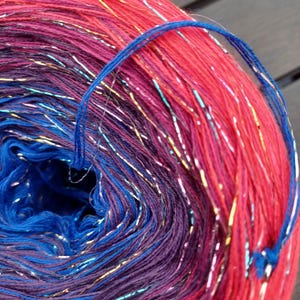 Könnte beinhalten: Nahaufnahme eines lebendigen Garnknäuels mit einem Farbverlauf aus Rot, Lila und Blau. Das Garn hat metallische Silber- und Goldfäden, und ein blauer Faden ist um das Knäuel geschlungen.