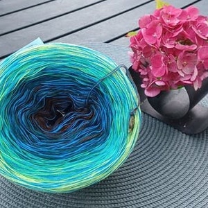 May include: A skein of yarn with a gradient of green, blue, and brown colors. The yarn is wound into a ball shape and is sitting on a gray woven mat. A bouquet of pink flowers is in the background.