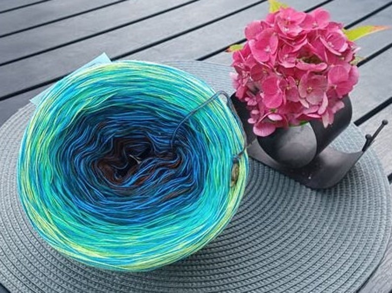 May include: A skein of yarn with a gradient of green, blue, and brown colors. The yarn is wound into a ball shape and is sitting on a gray woven mat. A bouquet of pink flowers is in the background.