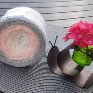 Könnte beinhalten: Ein großer Garnknäuel mit Farbverlauf in Weiß, Pfirsich und Grau liegt auf einem grauen Platzset. Eine dekorative Metallschnecke hält eine kleine grüne Glasvase mit rosa Blüten.