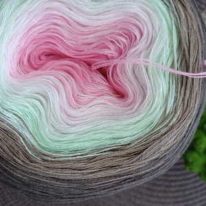 Könnte beinhalten: Eine Garnrolle mit einem Farbverlauf von Braun, Grün, Weiß und Rosa. Das Garn ist zu einer Kugel gewickelt.
