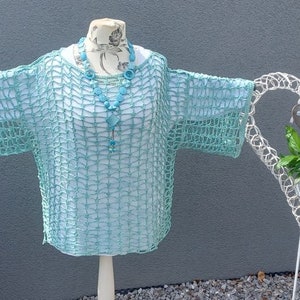 Könnte beinhalten: Ein hellblaues Häkeltop mit einer türkisfarbenen Perlenkette. Das Oberteil wird auf einem weißen Mannequin vor einem grauen Hintergrund präsentiert.