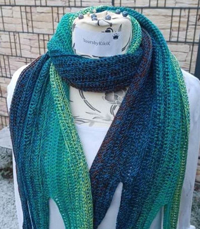 May include: A teal, green, and blue ombre crochet scarf with a brown stripe. The scarf is displayed on a white mannequin with the text "YoursbyKiki" on the label.