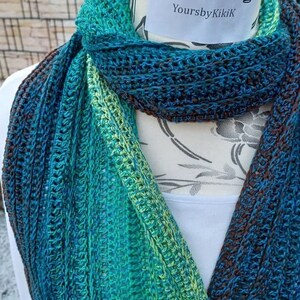 May include: A crocheted scarf in shades of teal, green, and brown. The scarf is displayed on a white mannequin with the text "YoursbyKikiK" on a white tag.
