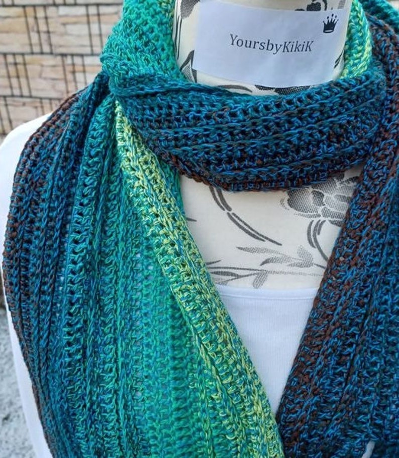 May include: A crocheted scarf in shades of teal, green, and brown. The scarf is displayed on a white mannequin with the text "YoursbyKikiK" on a white tag.