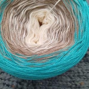 Könnte beinhalten: Eine Garnrolle mit einem Farbverlauf von Beige, Weiß und Türkis. Das Garn ist zu einer Kugel gewickelt.