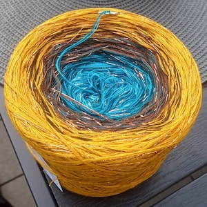 Könnte beinhalten: Eine Garnrolle mit einem Farbverlauf von Gelb, Braun und Blau. Das Garn hat einen metallischen Glanz.