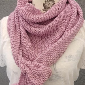 Könnte beinhalten: Ein gestrickter Schal in einem sanften Mauve-Ton, mit einem strukturierten Stich und zwei passenden Bommeln. Der Schal ist um ein weißes Oberteil drapiert und zeigt sein gemütliches Design. Der Schal hat eine dreieckige Form.