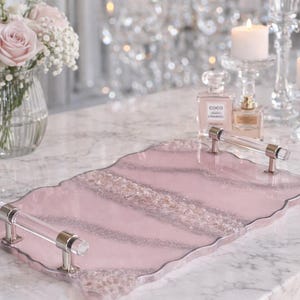 Puede incluir: Una bandeja de resina rosa con detalles de purpurina plateada y asas de acrílico transparente. La bandeja tiene un borde ondulado y se exhibe sobre una superficie de mármol. Es un artículo decorativo.