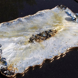 Peut inclure: Un plateau de service en résine blanche avec un bord pailleté or et des accents de paillettes noires et dorées. Le plateau a deux boutons en cristal sur les côtés.