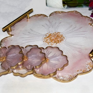 May include: A set of four pink and gold resin coasters with a flower design. The coasters are arranged in a stack, with the largest coaster on the bottom and the smallest coaster on top. The coasters have a gold rim and a flower design in the center. The coasters are perfect for protecting your furniture from scratches and spills.