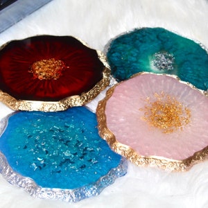 May include: Four agate coasters with gold trim. The coasters are in shades of red, teal, pink, and blue. Each coaster has a unique pattern of gold flecks.