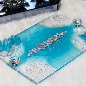 Puede incluir: Una bandeja rectangular con un diseño de resina azul y blanca. La bandeja tiene purpurina plateada incrustada en la resina. La bandeja tiene dos pomos de cristal en los lados.