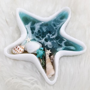 May include: A white, star-shaped resin dish with a turquoise and white wave design. The dish is filled with seashells, a small turquoise turtle figurine, and tiny white starfish. The dish is on a white, fluffy surface.