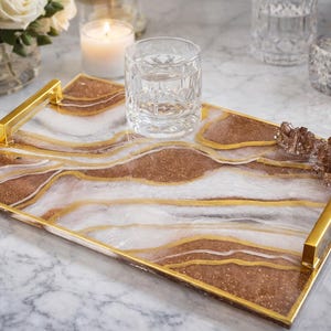 May include: A rectangular serving tray with gold handles and trim. The tray features a brown, white, and gold marbled design. A clear glass of water sits on the tray. Decorative items are in the background.