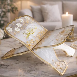 May include: An ornate, open book-shaped stand with a gold trim, white floral accents, and gold leaf details. The stand displays Arabic script in gold. A string of pearls with a gold tassel rests nearby. The stand is on a marble table.