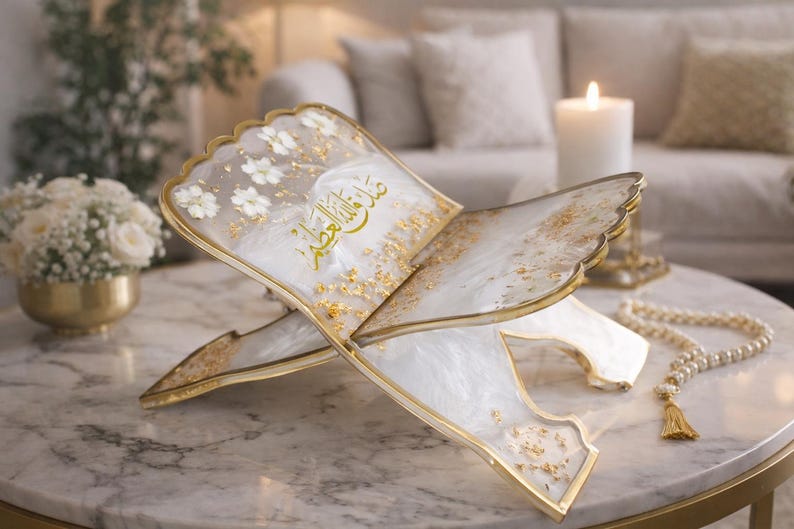 May include: An ornate, open book-shaped stand with a gold trim, white floral accents, and gold leaf details. The stand displays Arabic script in gold. A string of pearls with a gold tassel rests nearby. The stand is on a marble table.