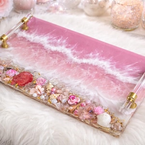 Könnte beinhalten: Ein rechteckiges Serviertablett mit einem rosa-weißen Wellendesign, verziert mit Muscheln, Blumen und goldenen Akzenten. Das Tablett hat transparente Acrylgriffe mit goldenen Beschlägen. Das Thema ist maritim und dekorativ.