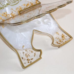 May include: A decorative, clear resin book stand with gold leaf accents. The stand has a white, pearlescent base and gold trim. The top edge has a scalloped design. Gold Arabic script is visible on the top.