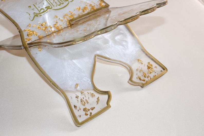 May include: A decorative, clear resin book stand with gold leaf accents. The stand has a white, pearlescent base and gold trim. The top edge has a scalloped design. Gold Arabic script is visible on the top.