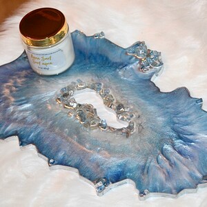 May include: A decorative resin tray in shades of blue and silver, with a geode-like design and clear crystal accents. A jar with a gold lid and a label that reads "Aqua Surf sweet aqua" sits on the tray. The tray is approximately 10 inches long.