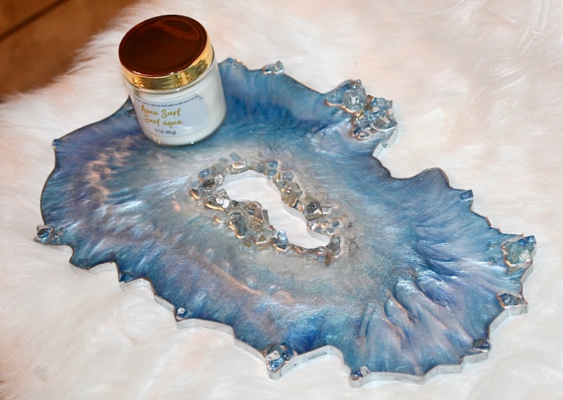 May include: A decorative resin tray in shades of blue and silver, with a geode-like design and clear crystal accents. A jar with a gold lid and a label that reads "Aqua Surf sweet aqua" sits on the tray. The tray is approximately 10 inches long.
