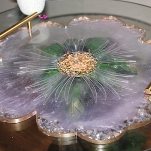 May include: A light purple resin serving tray with gold accents and a flower design. The tray has two gold handles and a clear center with gold flakes.