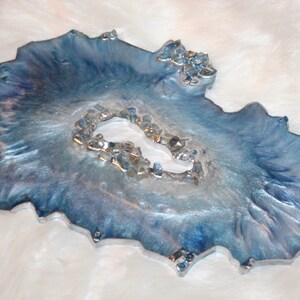 May include: A decorative resin tray with a geode-like design. The tray features shades of blue and silver with a textured surface and embedded crystals. The tray has an irregular, organic shape, and is approximately 10 inches in length.