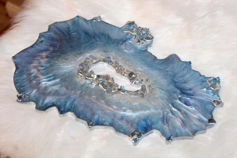 May include: A decorative resin tray with a geode-like design. The tray features shades of blue and silver with a textured surface and embedded crystals. The tray has an irregular, organic shape, and is approximately 10 inches in length.
