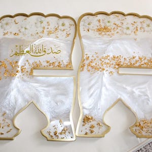 May include: Two decorative resin pieces with a white and gold design. The pieces have a unique shape with a gold border and are filled with white resin, gold flakes, and small floral accents. Arabic text is visible.