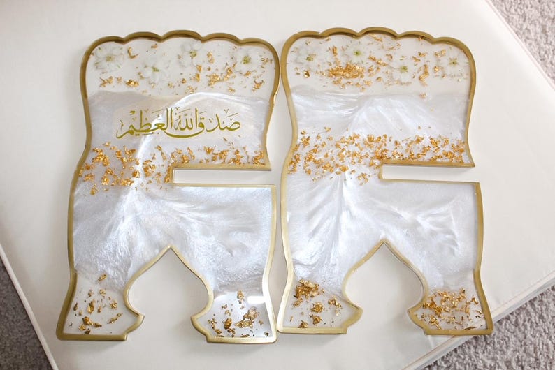 May include: Two decorative resin pieces with a white and gold design. The pieces have a unique shape with a gold border and are filled with white resin, gold flakes, and small floral accents. Arabic text is visible.