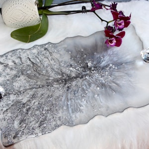 May include: A silver and white resin tray with a glittery finish and a unique, organic shape. The tray has two crystal knobs on the sides.