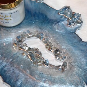 May include: A decorative resin tray with a blue and silver marbled design, accented with embedded glass pieces. A white candle with a gold lid and the text "Aqua Surf Surf aqua" is visible. The candle weighs 3 oz (85g).