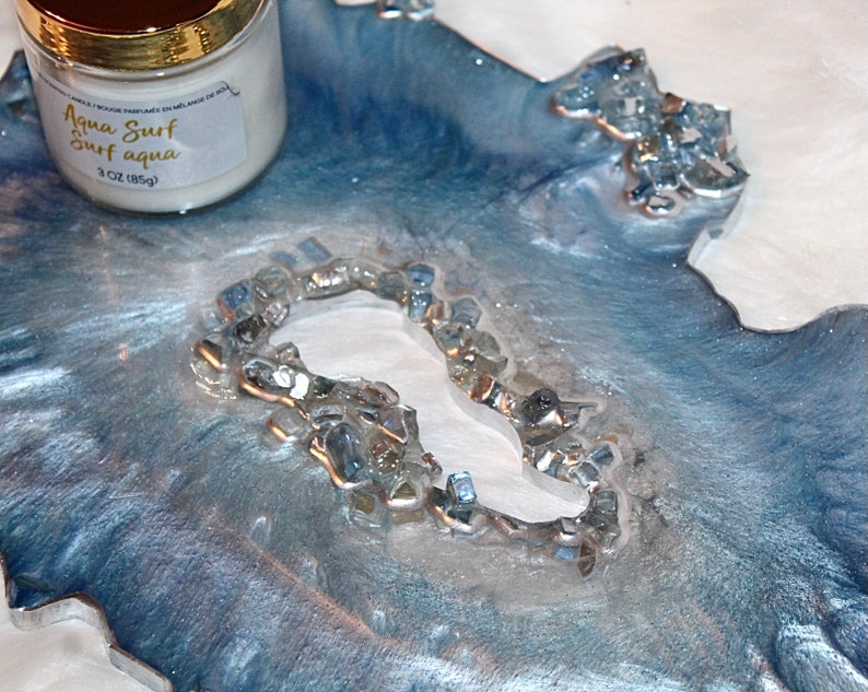 May include: A decorative resin tray with a blue and silver marbled design, accented with embedded glass pieces. A white candle with a gold lid and the text "Aqua Surf Surf aqua" is visible. The candle weighs 3 oz (85g).