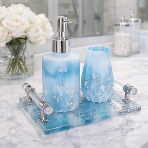 Puede incluir: Un juego de baño con dispensador de jabón, vaso y bandeja. Los artículos son de color azul claro con detalles plateados. La bandeja tiene asas transparentes. Un jarrón de rosas blancas se encuentra en el fondo.