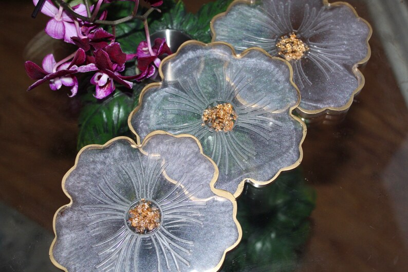 May include: Three clear resin flower-shaped coasters with gold trim and gold glitter accents.