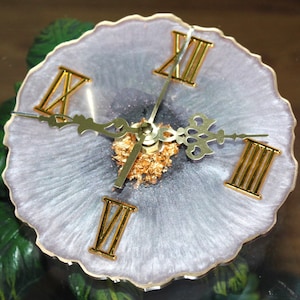 May include: A white and silver resin clock with gold Roman numeral hour markers and gold hands. The clock face is embedded with gold flakes.