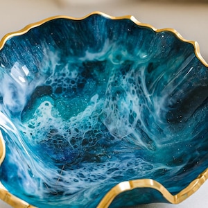 Peut inclure: Bol décoratif avec un bord ondulé de couleur or. L'intérieur présente un motif tourbillonnant de résine bleue et blanche, ressemblant à des vagues océaniques. Les couleurs du bol varient du bleu marine profond au turquoise clair.