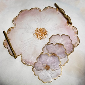 May include: A set of four pink and white resin coasters with a floral design. The coasters are shaped like flowers and have gold accents. The coasters are on a white surface.