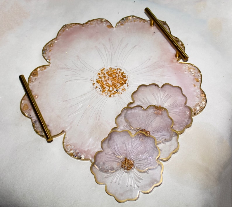 May include: A set of four pink and white resin coasters with a floral design. The coasters are shaped like flowers and have gold accents. The coasters are on a white surface.