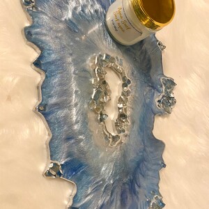 May include: A decorative resin tray with a blue and silver marbled design. The tray has an irregular shape and is embellished with silver-colored stones. A white jar with gold trim and the text "Aqua Surf Surf Aqua" is on the tray.