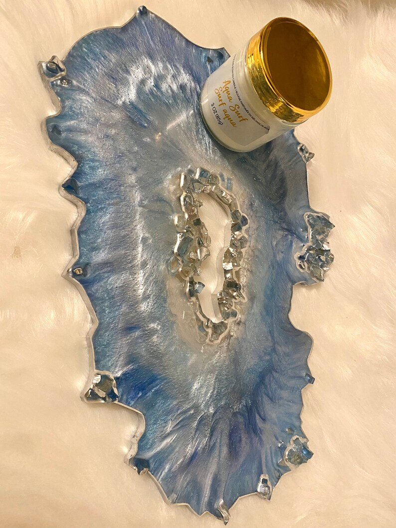 May include: A decorative resin tray with a blue and silver marbled design. The tray has an irregular shape and is embellished with silver-colored stones. A white jar with gold trim and the text "Aqua Surf Surf Aqua" is on the tray.