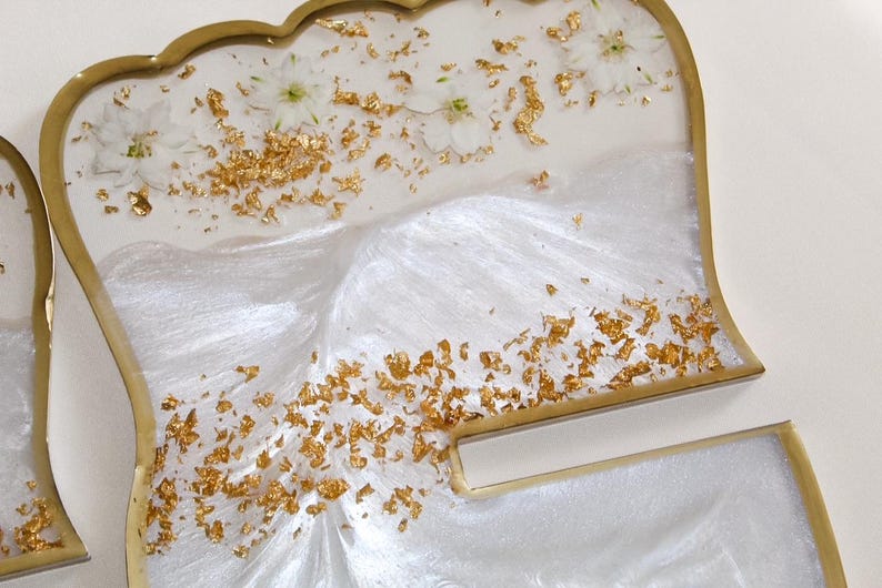 May include: Decorative resin trays with a scalloped edge and gold trim. The trays are white with gold leaf accents and small white flowers embedded in the resin. The design has a pearlescent, shimmering effect.