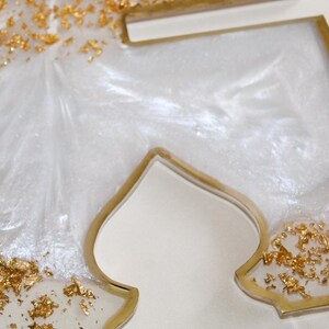 May include: Close-up of a decorative resin piece in a stylized shape. The piece features a white, pearlescent interior with gold leaf accents. The edges are outlined in gold, creating a luxurious and elegant aesthetic.