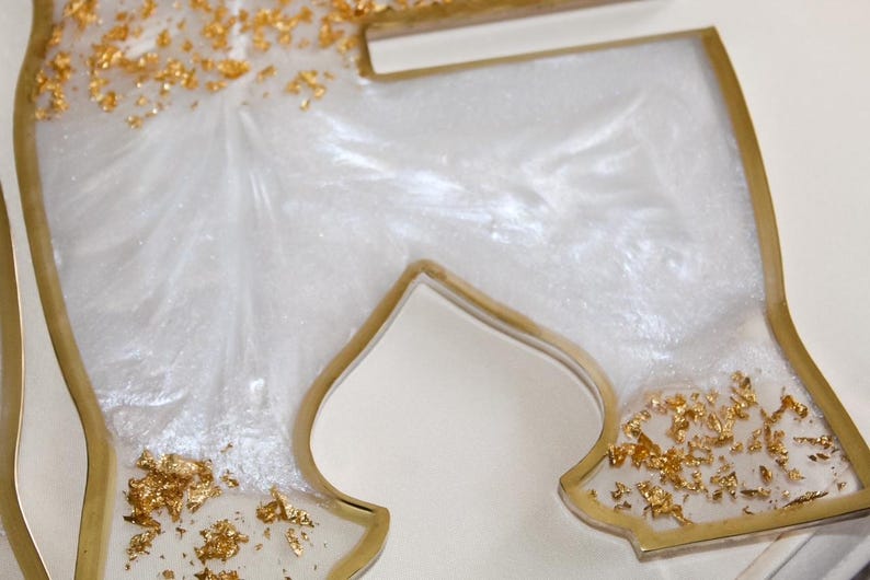 May include: Close-up of a decorative resin piece in a stylized shape. The piece features a white, pearlescent interior with gold leaf accents. The edges are outlined in gold, creating a luxurious and elegant aesthetic.