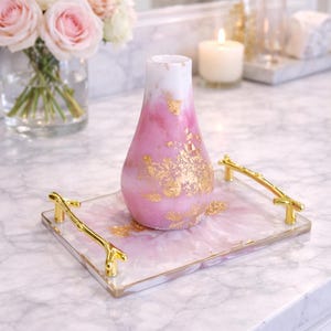 May include: A pink and white decorative vase with gold leaf accents sits on a rectangular tray. The tray has gold handles and a pink and white marbled design. A bouquet of pink roses is in the background.